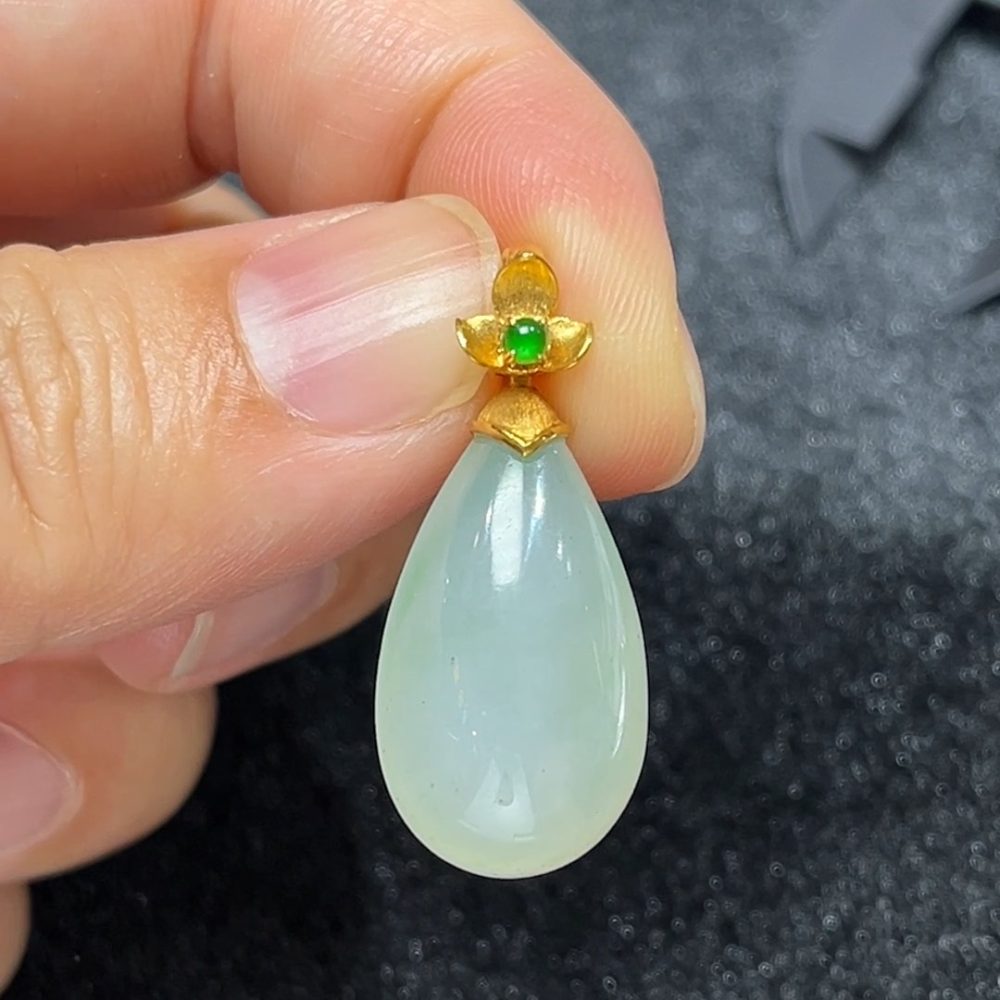這款天然A級翡翠18K金鑲嵌水滴吊墜（編號#299），採用色澤清新柔和的淺綠色翡翠精工雕琢，水滴造型線條圓潤流暢，如露珠凝翠、靜靜垂掛胸前，展現自然與靈動之美。翡翠質地通透溫潤，飽含東方玉石的靈性與氣韻；外圈採用18K金鑲嵌設計，細膩包邊，不僅提升整體質感，更增添了一抹現代奢雅氣息。此款設計極具女性柔美氣質，是日常佩戴或優雅禮贈的上乘之選。
